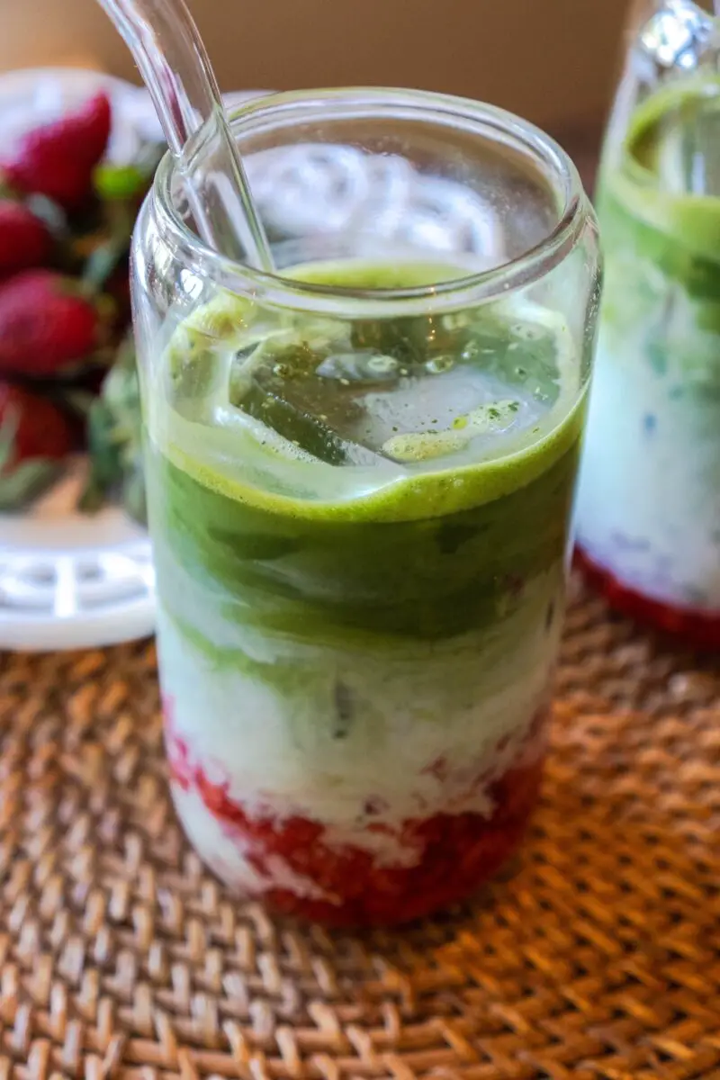 A photo of a strawberry matcha latte in a glass with a glass straw. Strawberries sit in the background. 