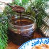 Bourbon Sauce in a jar next to greenery and a plate of pretzels. A spoon drizzling chocolate sauce back into jar