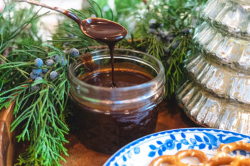 Bourbon Sauce in a jar next to greenery and a plate of pretzels. A spoon drizzling chocolate sauce back into jar