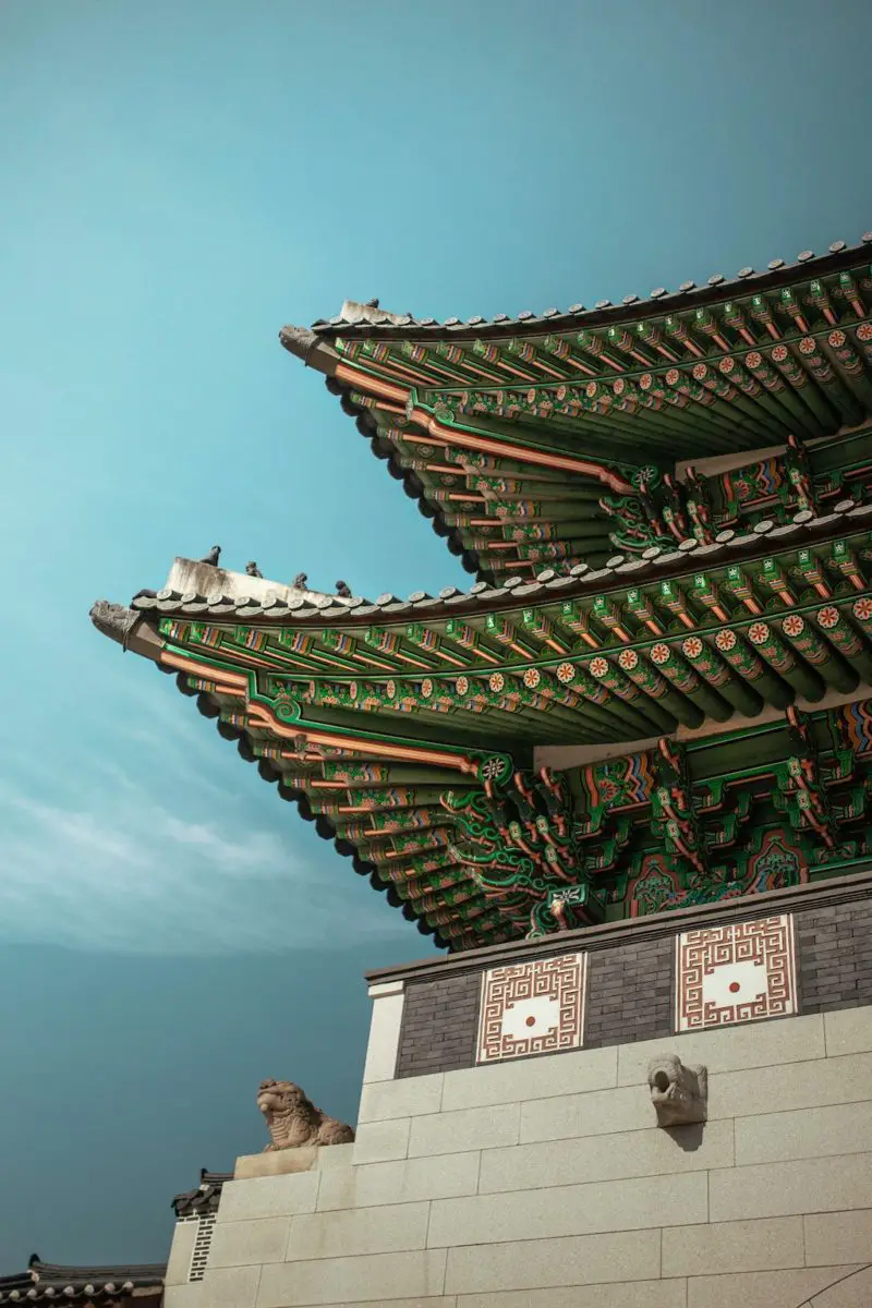 A look at a traditional Korean building. The building sits high up. The underside of the roof is filled will different colors and designs. 