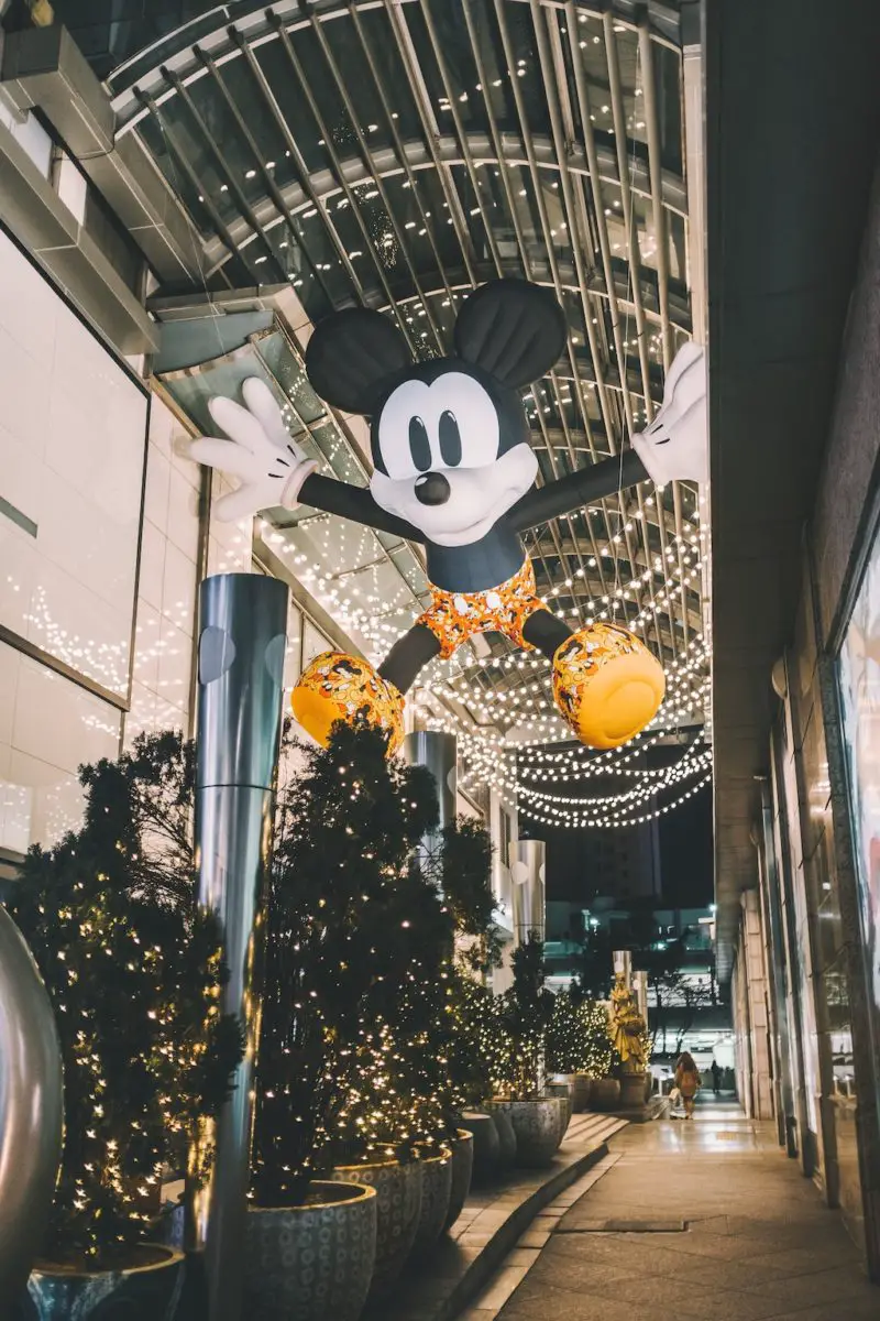 Holiday decorations in an alley in Seoul, South Korea. Bushes are lit with lights, strings of lights are overhead, and a giant Mickey Mouse blow up doll in hanging overhead.