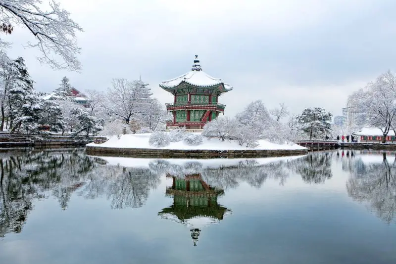 A winter photo of a pavilion that sits in the center of Gyeongbokgung Palace in Seoul, Korea. The pavilion sits in the middle of a pond covered in snow. 