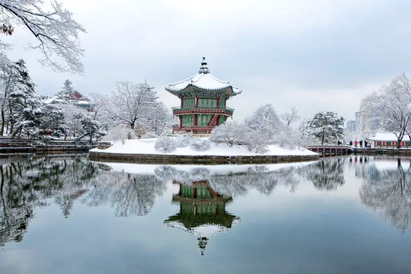 A winter photo of a pavilion that sits in the center of Gyeongbokgung Palace in Seoul, Korea. The pavilion sits in the middle of a pond covered in snow.