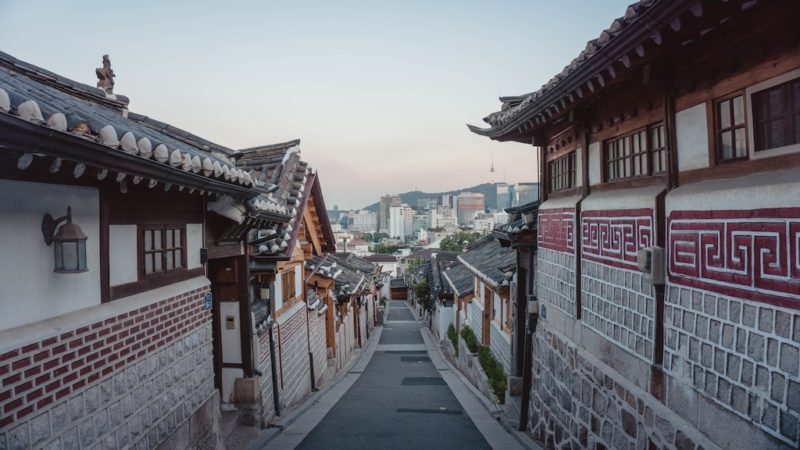 A traditional Korean street--a narrow road with old houses on the side. The houses are white with brown wood. The roofs are slate. 