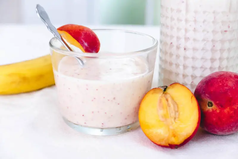 A close up shot of banana peach smoothie--peaches sit next to the smoothie. 