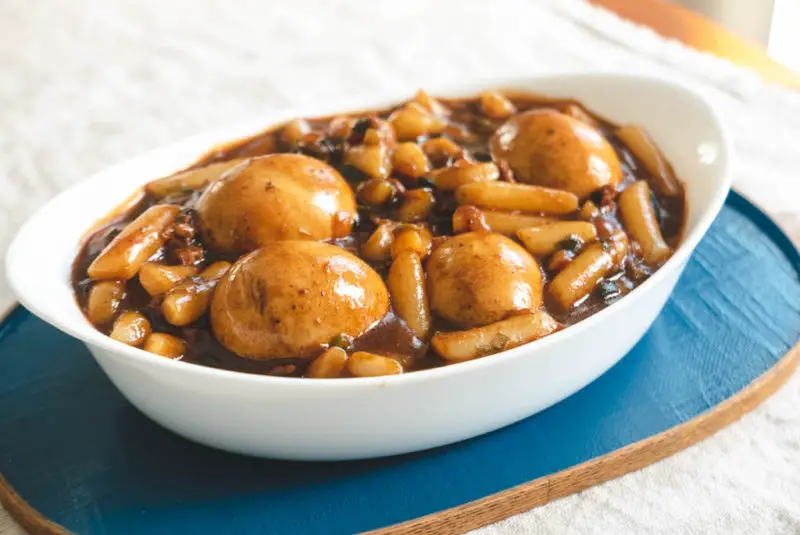 Korean jajang tteokbokki-- a white bowl on a blue tray. The bowl contains rice cakes in a brown sauce with eggs. 