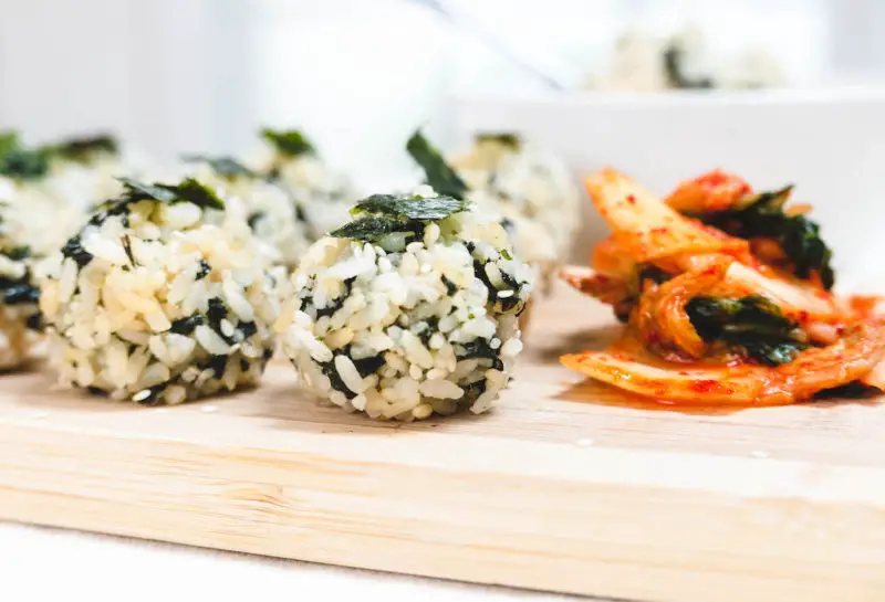 A closeup shot of South Korean vegan handmade rice balls. The rice is shaped into balls filled with dried seaweed, soy sauce, and more. It sits next to kimchi.