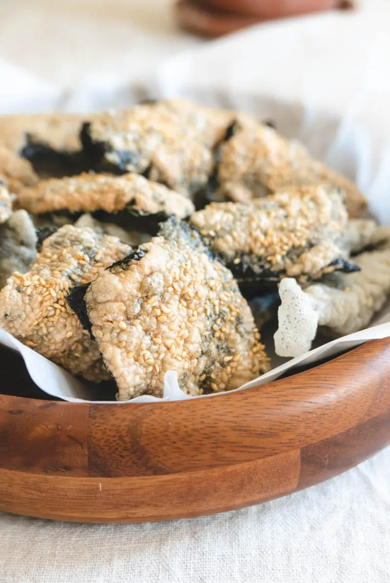 A closeup side shot of a wooden bowl with traditional Korean vegan fried seaweed snacks on the inside. The snacks have sesame seeds on top.