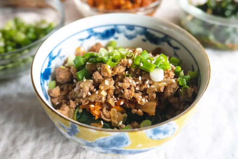 an overheat side shot of a Korean grits bowl--green onions, ground pork, kimchi, spinach on grits.