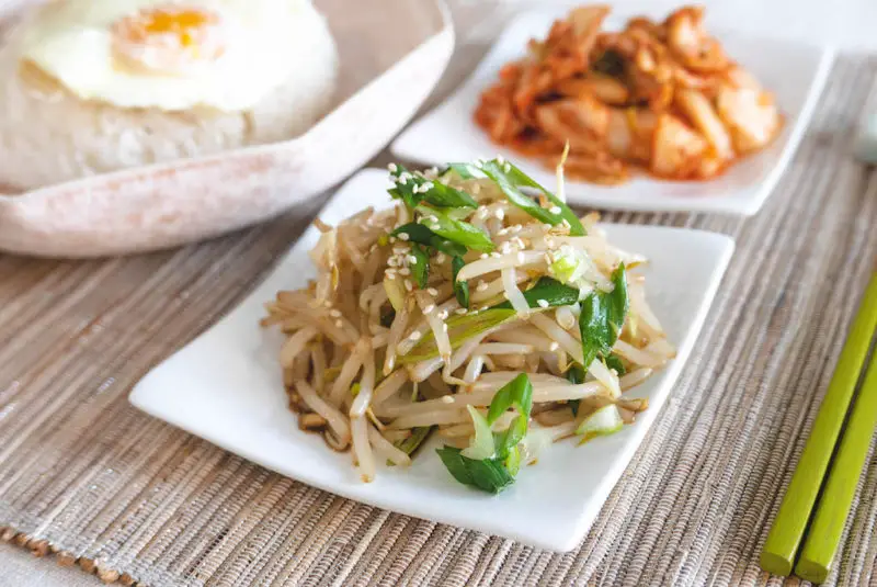 A side shot of sukju-namul muchim next to bright red kimchi, chopsticks, and a bowl of rice with eggs. 