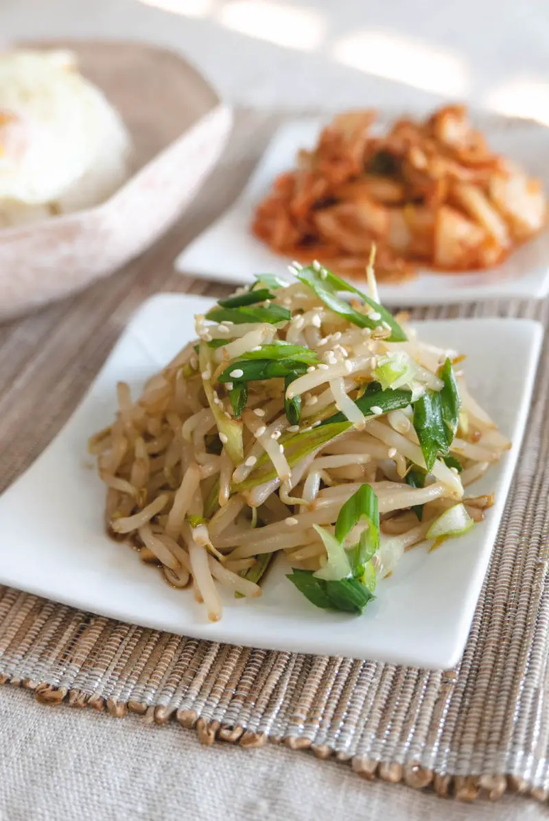 A close up side shot of mung bean sprout salad. The salad is topped with green onions and sesame seeds. Kimchi sits behind the salad.