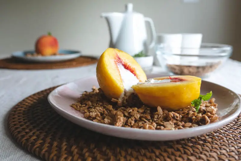 A peach with greek yogurt on the inside sliced in half. Granola sits on the plate around the peach. 