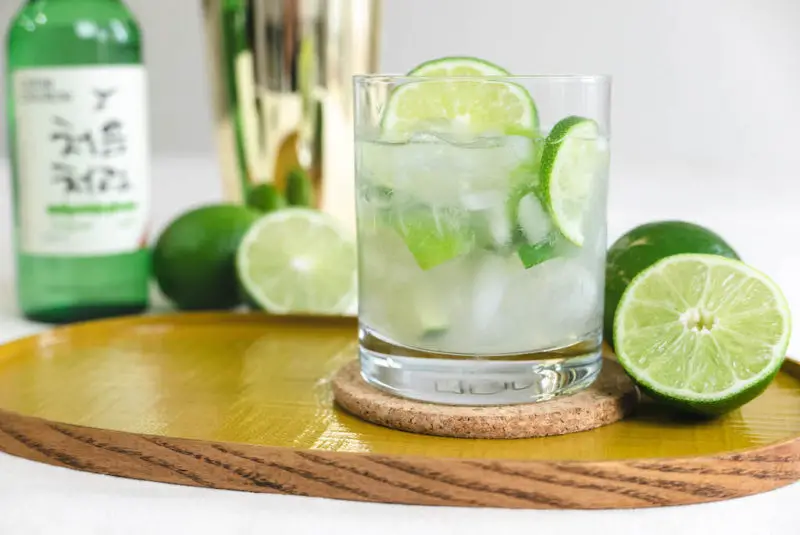 A glass of soju caipirinha on a yellow tray. limes sit next to the glass and a bottle of soju sits in the background. 