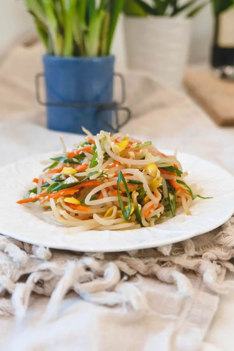 A side shot of kongnamul muchim on a white plate. The plate sits on a linen table cloth with plants in the background.