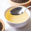 A spoonful of perilla oil hovering over a white bowl filled with perilla oil. Seeds sit around the bowl of oil.