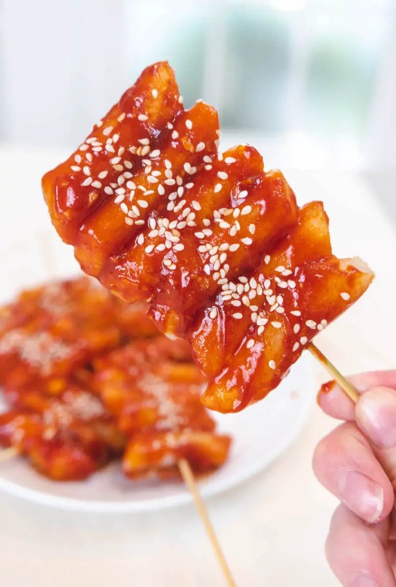 A side shot of bright red Korean rice cake skewers topped with sesame seeds.