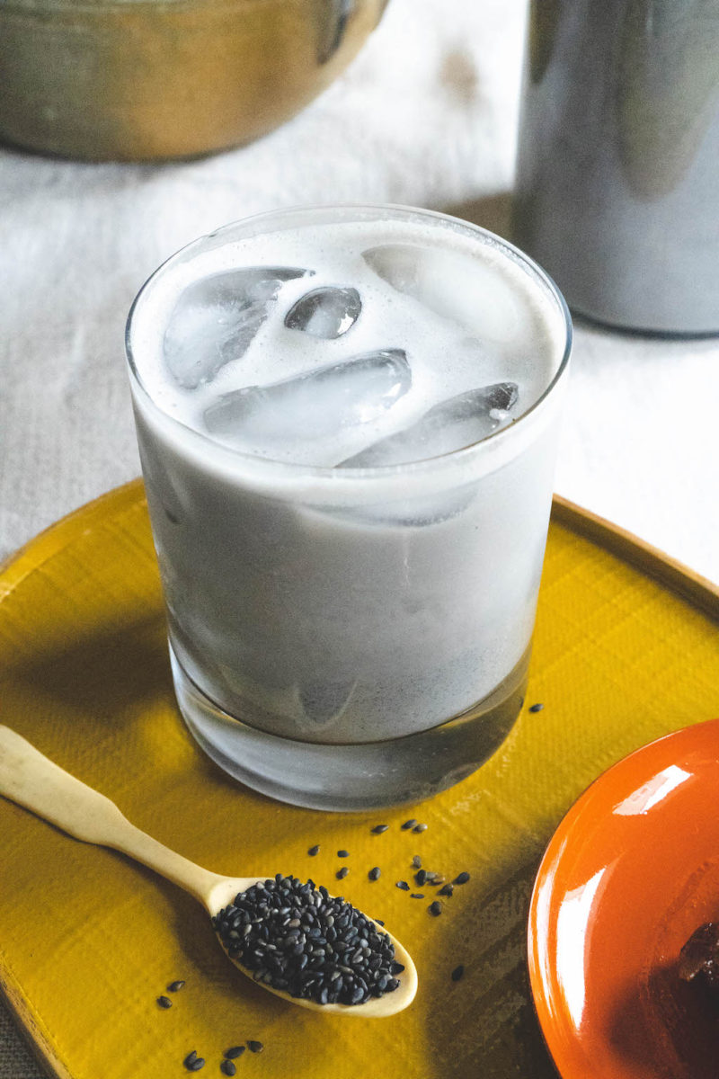 A overhead side shot of black sesame milk on a yellow tray. Black sesame seeds sit next to the cup of milk on a spoon.