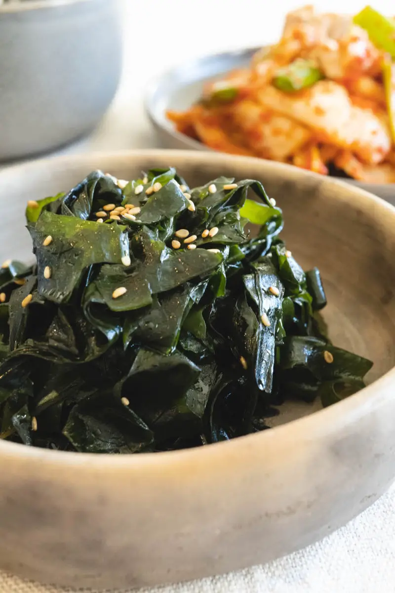 A side shot of miyeok namul in a stone bowl. Red fresh kimchi sits behind the bowl. 