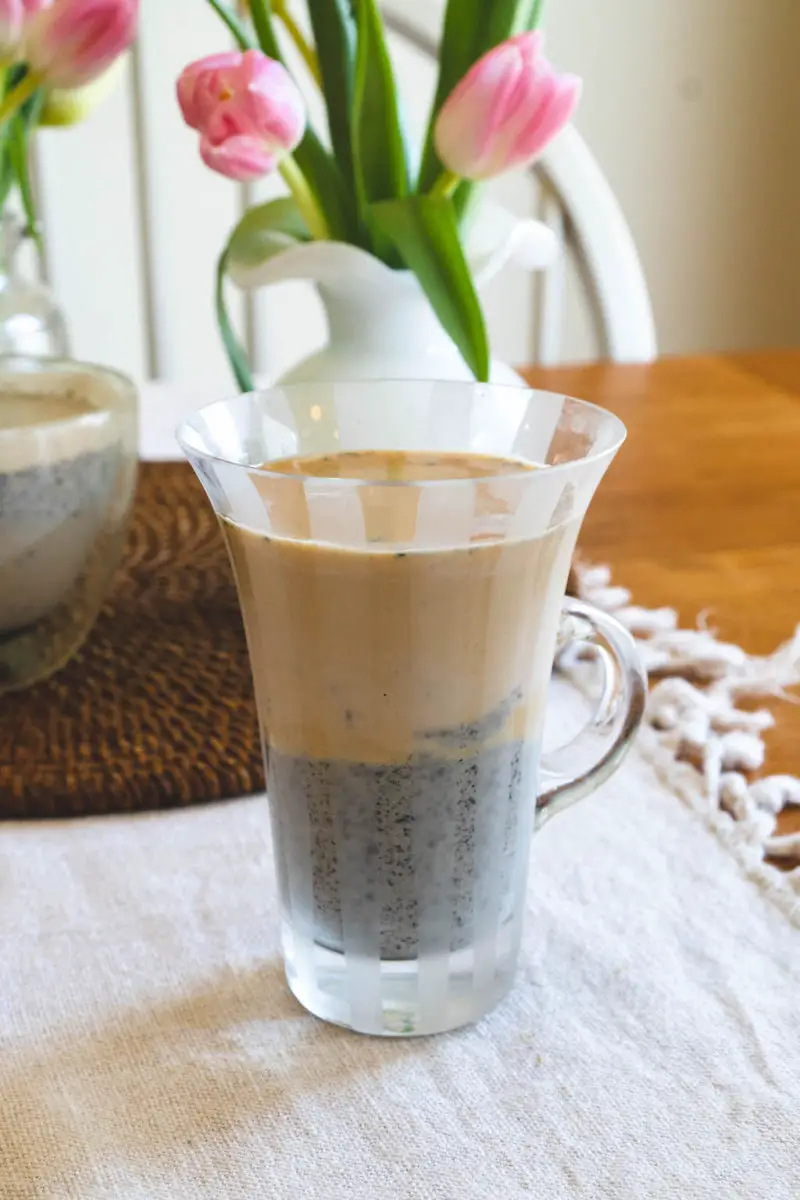 A side shot of a black sesame latte next to flowers. The latte has a bottom layer of black sesame paste and top layer contains milk and espresso.