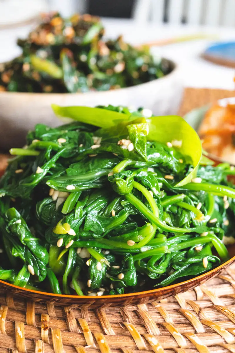 A side shot of Korean spinach side dish on a wicker mat. Other vegetable side dishes sit behind it. 
