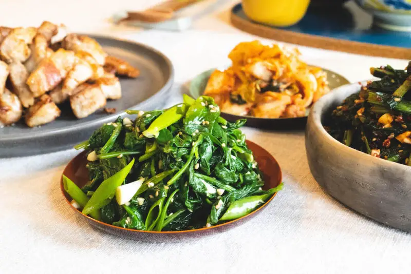 A side shot of Korean spinach side dish next to grilled pork belly, kimchi, and dandelion greens salad. 