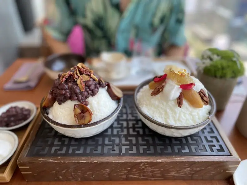 Two bowls of bingsu on a tray. The left is patbingsu topped with red beans and jujubes. The right is topped with honeycomb. 