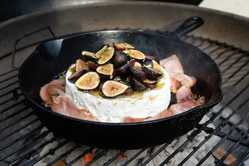 Brie being cooked on the campfire in a cast iron skillet topped with figs, fig jam, prosciutto, and rosemary.