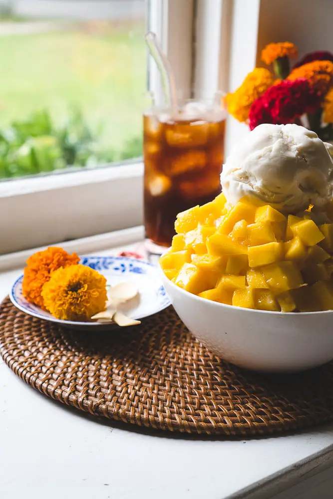 A bowl of Korean mango shaved ice next to a plate with spoons and iced coffee.