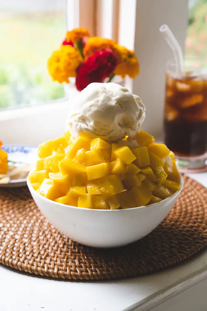 A closeup side shot of Korean mango shaved ice. Flowers and coffee sit behind. 