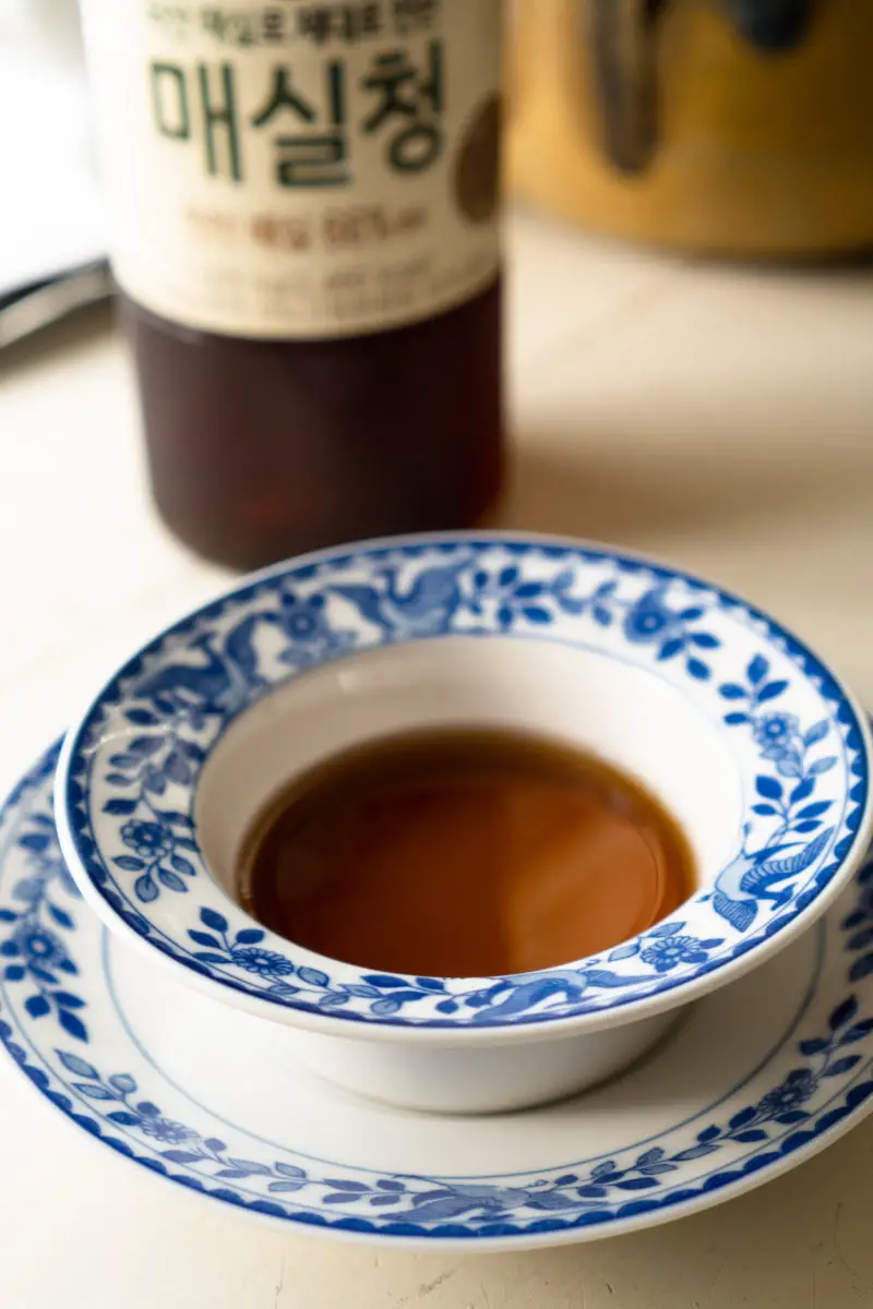 A side shot of Korean green plum syrup in a blue and white bowl. The bottle of the syrup sits behind. 