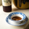 Maesil cheong in a blue and white bowl sitting on a window sill. The bottle of Korean green plum syrup behind it.