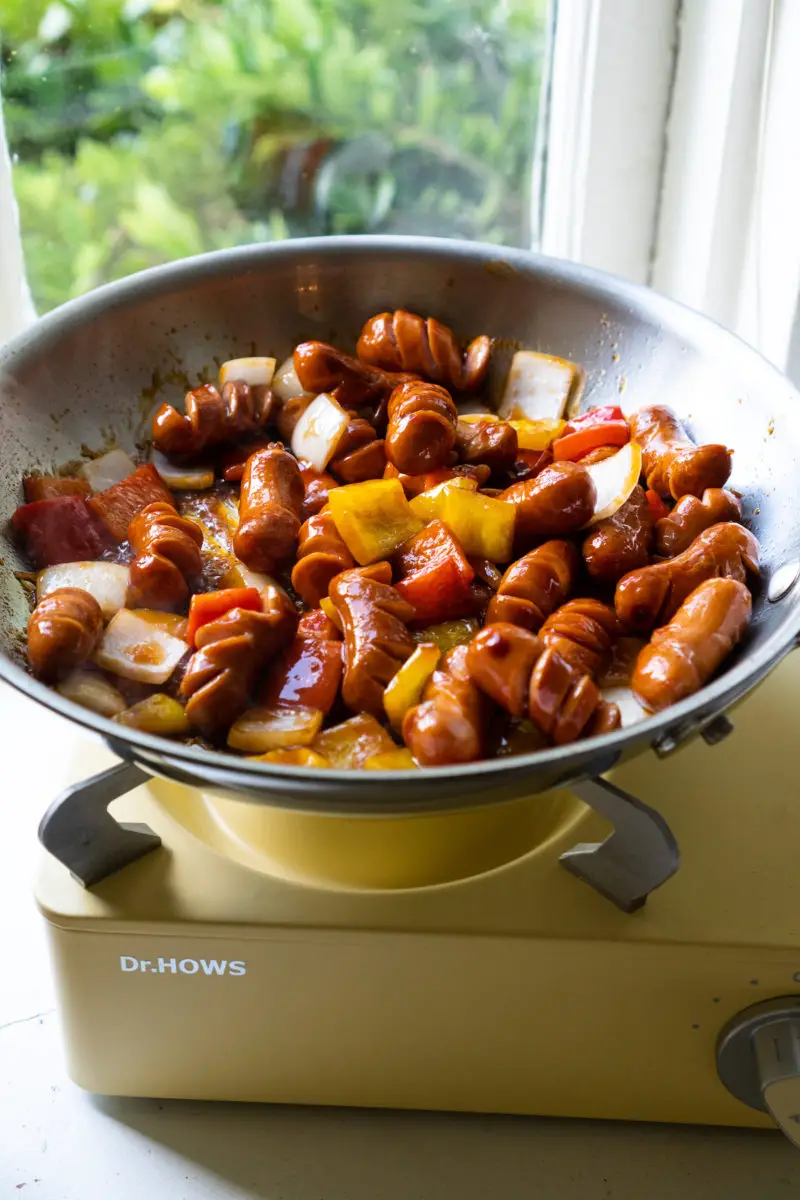 Sausage, onions, and peppers cooked on a yellow stovetop mixed with a thick sauce.