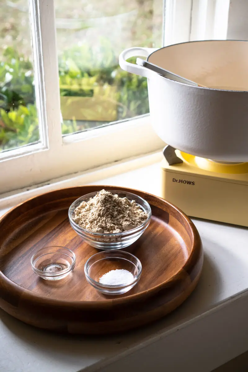 On a wooden tray, three bowls are set up. The bowls contain acorn starch, sesame oil, and salt. In the background is a gas stove and a white pot. 