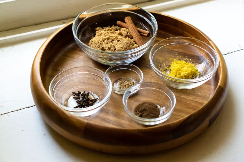 All the ingredients used to make spiced simple syrup in clear glass bowls. These include cinnamon, brown sugar, lemon zest, whole cloves, ground allspice, and ground coriander. 