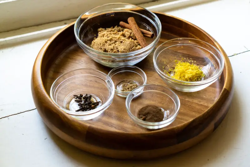 All the ingredients used to make spiced simple syrup in clear glass bowls. These include cinnamon, brown sugar, lemon zest, whole cloves, ground allspice, and ground coriander.