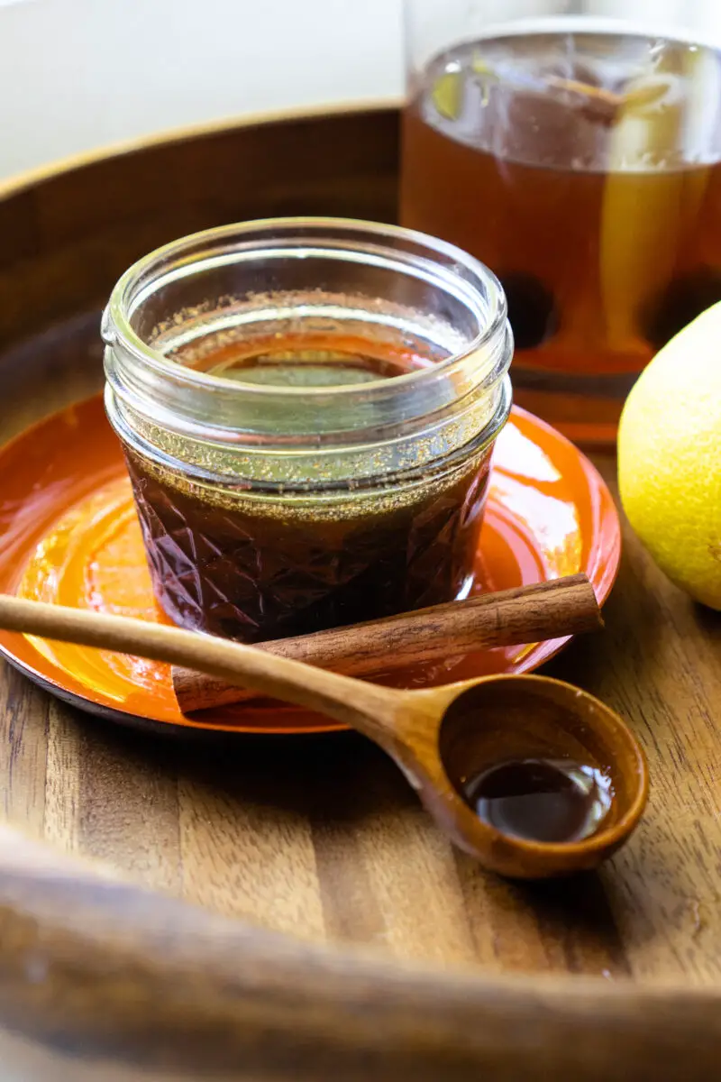A side shot of spiced simple syrup in a glass jar. It is sitting next to a spoon, lemon, and cinnamon stick. A drink sits in the background. 