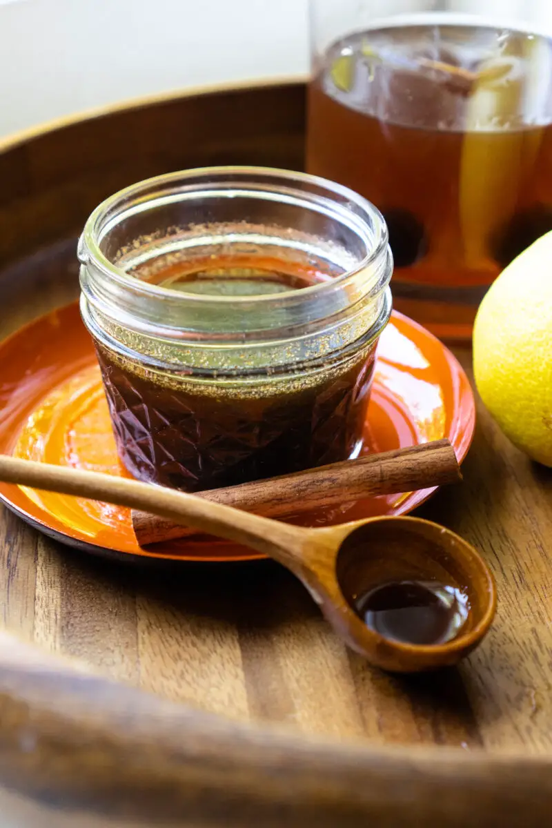 A side shot of spiced simple syrup in a glass jar. It is sitting next to a spoon, lemon, and cinnamon stick. A drink sits in the background.