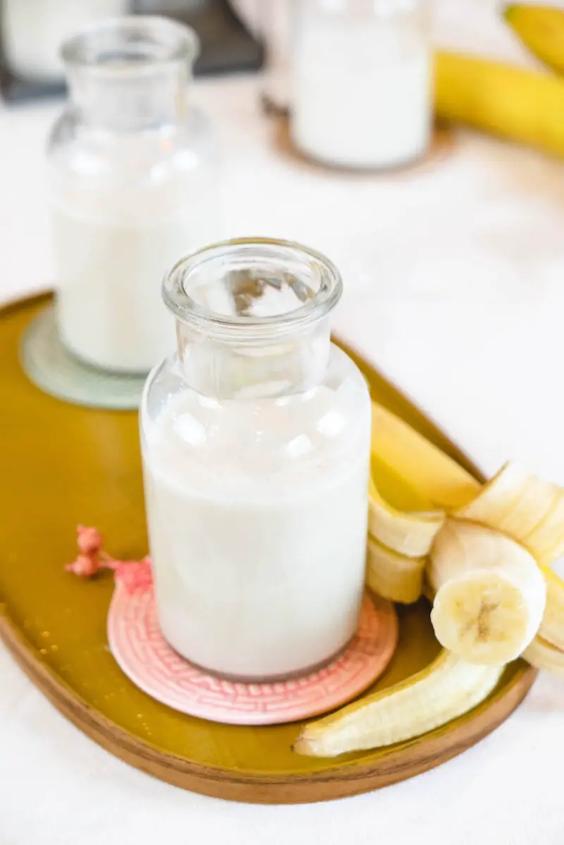 A side shot of banana-flavored milk sitting next to a banana on a yellow tray.