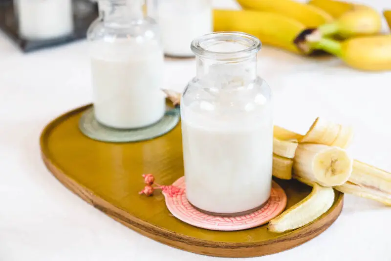 A side shot of two bottles of Korean banana milk on a yellow tray. A peeled banana (still in the peel) sits on the tray with the bottles. 