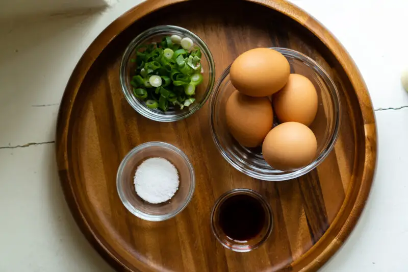 An overhead shot of the gyeran guk ingredients on a wooden tray. The ingredients include 4 eggs, chopped green onions, soy sauce, and salt. 