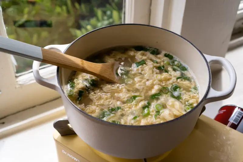 A side shot of Korean egg soup  with green onions in a white pot on yellow cooktop. 