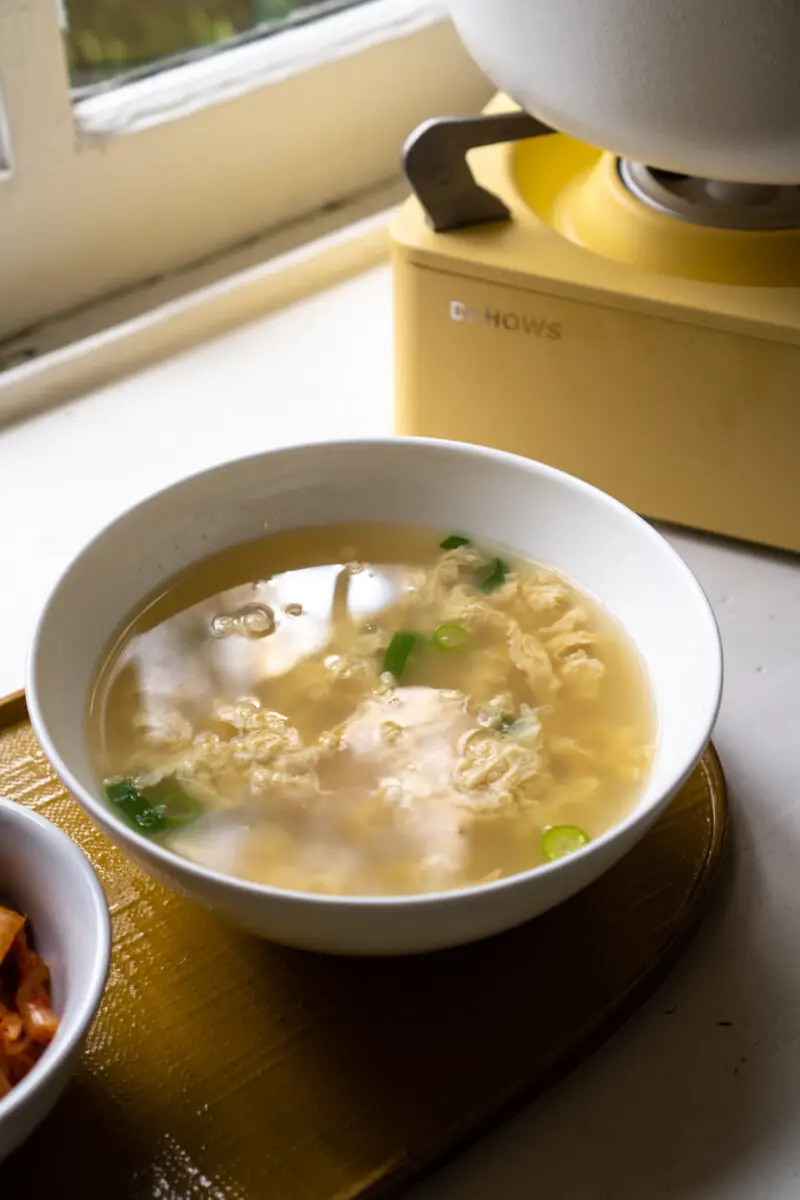 A side shot of Korea egg soup in a white bowl on a yellow tray.