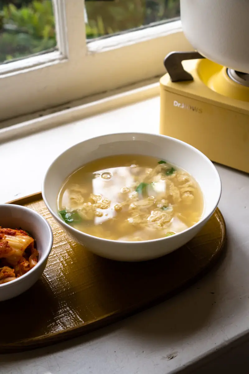 A side shot of Korean egg soup (gyeran guk) on a yellow tray next to kimchi. The yellow stovetop and white pot used to cook this dish sit in the background.