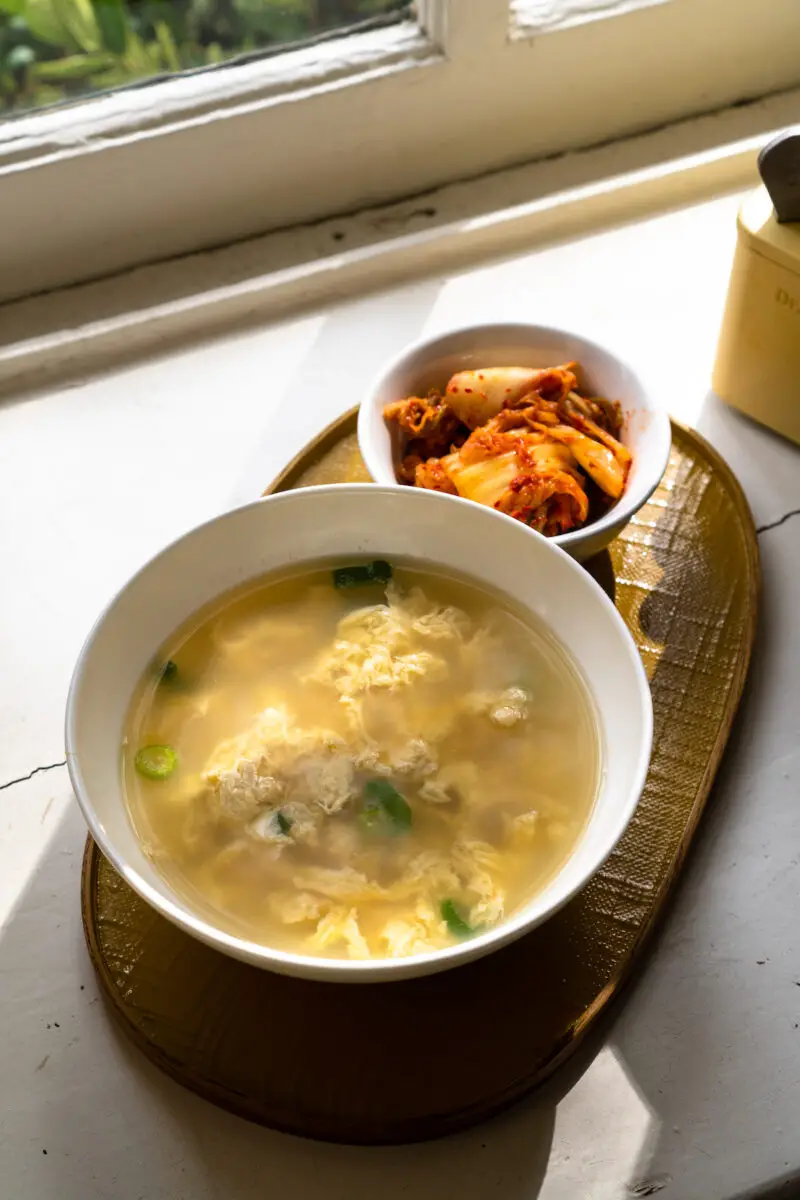 An overhead shot of a bowl of Korean egg soup (gyeran guk) in a white bowl. The bowl sits on a tray with kimchi.
