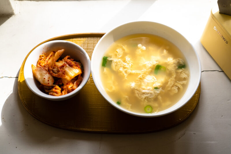 Korean egg soup on a yellow tray next to a bowl of kimchi. 