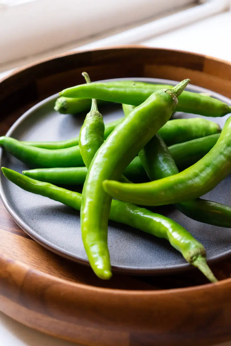 A side shot of a pile of Korean peppers (green) on a gray plate. The plate is sitting on a round wooden tray. 