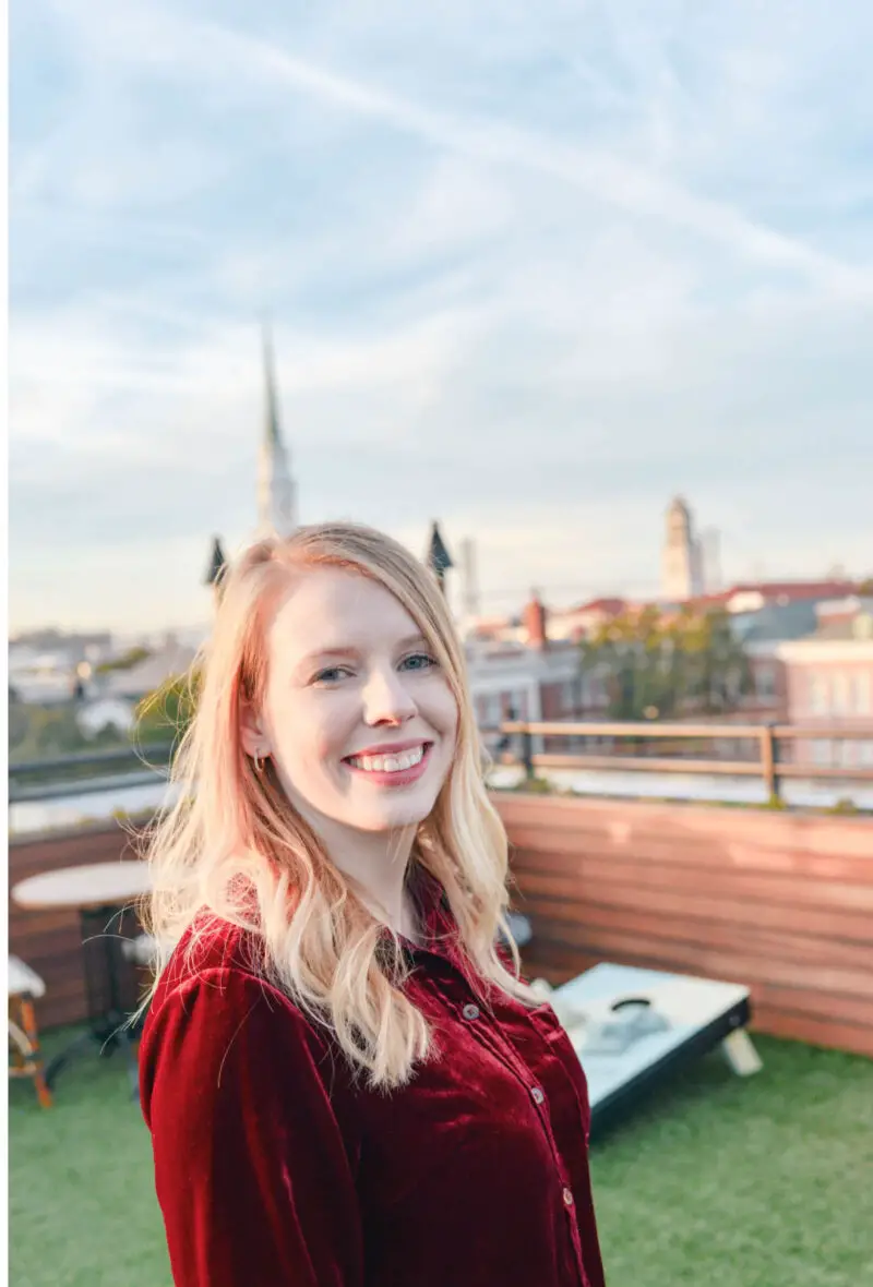 An image of the Emily, the owner of Carving A Journey. Emily is sideways but her face is facing toward the camera and she is smiling. She is wearing a red velvet top. She has blond hair and blue eyes. Behind her is the skyline of Savannah, Georgia. 