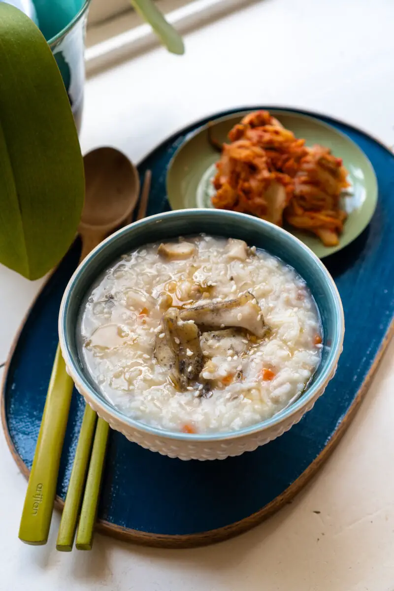 An overhead shot of Korean abalone porridge in a dotted bowl on a blue tray. Kimchi sits behind the tray and a wooden spoon and chopsticks sit next to the bowl.