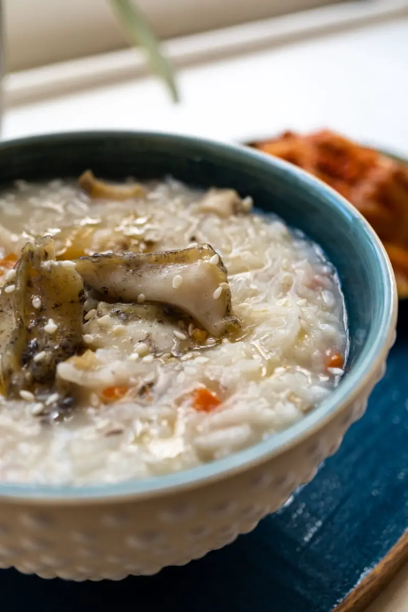 Korean abalone porridge (jeokbokjuk) in a dotted bowl with a blue interior. The bowl sits on a blue tray with kimchi behind it. 