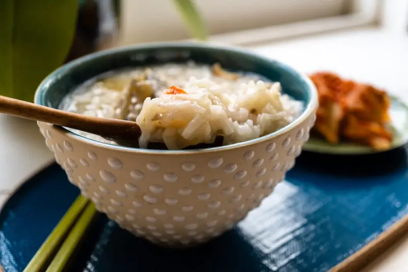 A side shot of jeonbokjuk in a dotted bowl on a blue tray. Kimchi sits behind the tray. A wooden spoon sits in the dish and is scooping some of it out. 